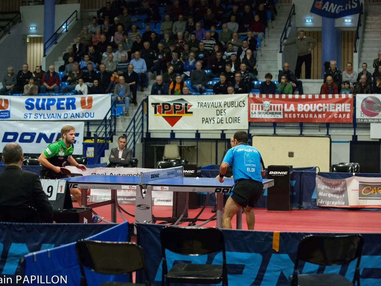 This picture shows a couple of men playing table tennis and we see couple of them watching by...