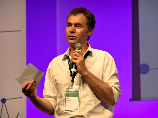 In the image there is a man in cream shirt and id card talking on mic holding a paper and behind...