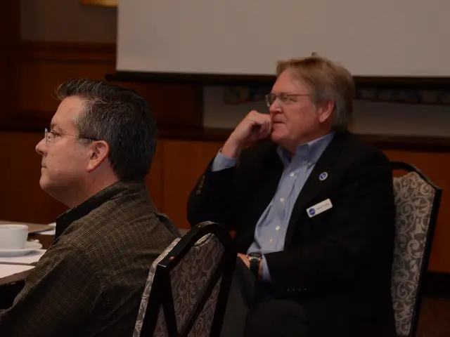 In this image I can see two men are sitting on chairs and I can see both of them are wearing specs....